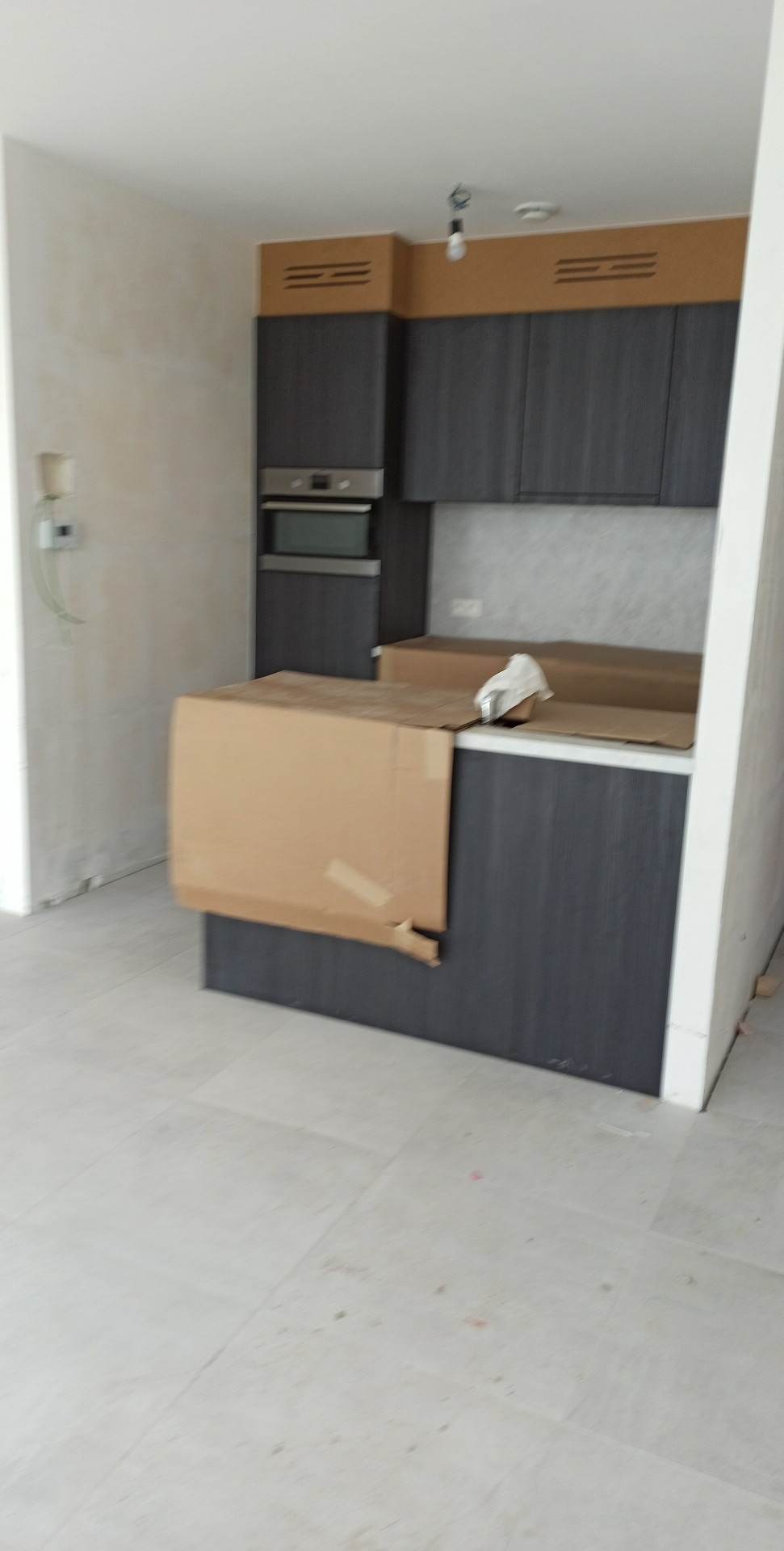 Wnętrze kuchni w trakcie wykańczania: zamontowane szare szafki kuchenne z blatem częściowo zakrytym kartonem ochronnym, surowe ściany w tle i podłoga z jasnych płytek.
