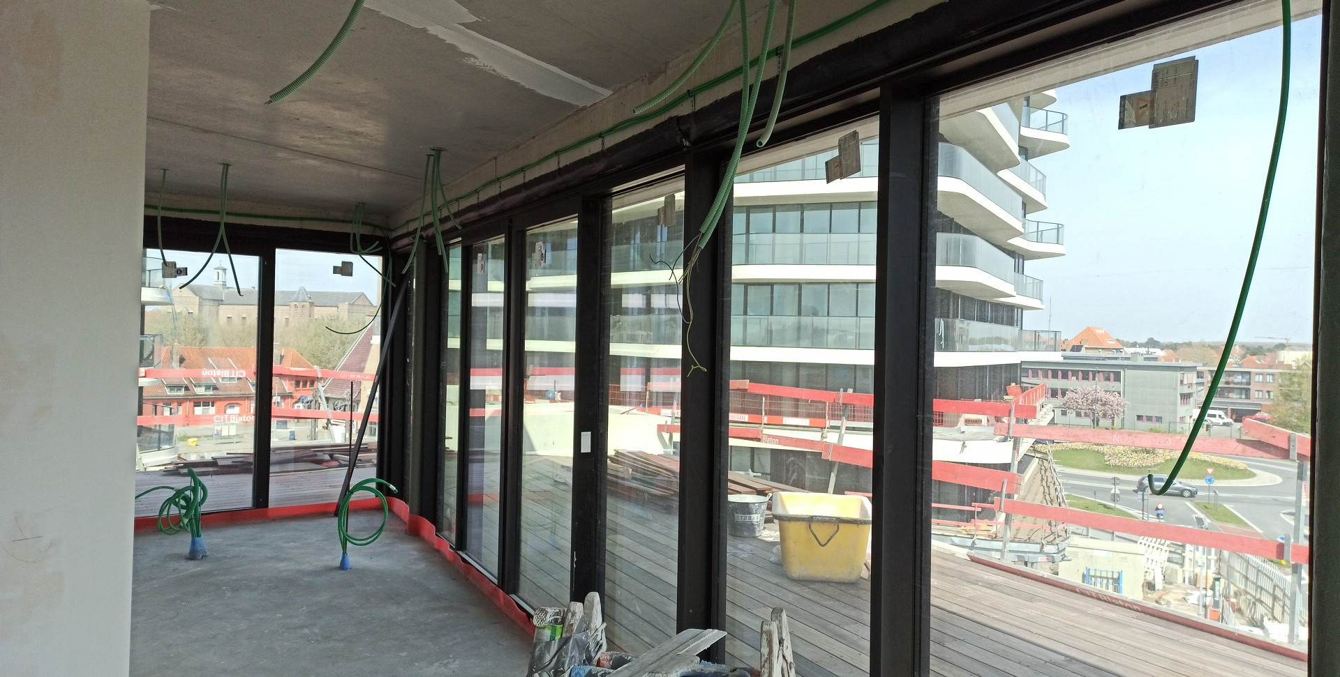 Wnętrze w trakcie wykańczania z dużymi oknami balkonowymi, widok na nowoczesny budynek z balkonami, widoczne przewody elektryczne pod sufitem i żółte wiadro.