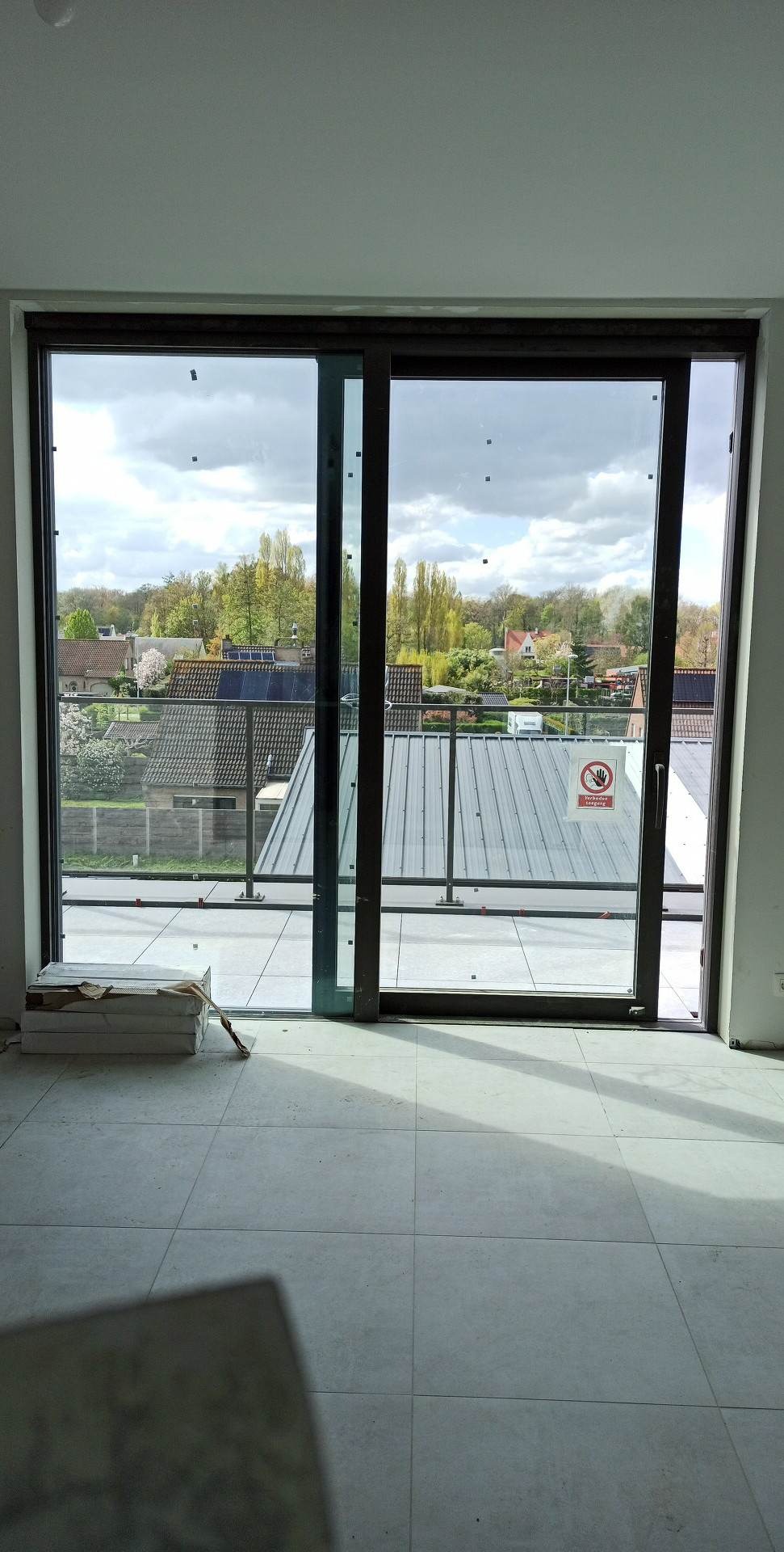 Wnętrze z dużym przesuwnym oknem balkonowym, widok na zewnątrz z metalową balustradą i dachami domów, na podłodze ułożone jasne płytki, w rogu paczka płytek.