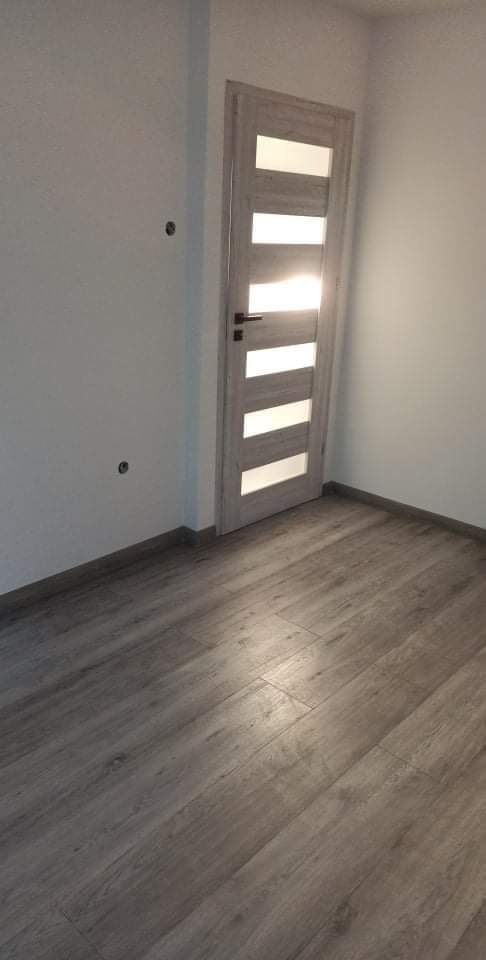 Wykończone wnętrze z panelami podłogowymi imitującymi drewno i nowoczesnymi drzwiami z przeszkleniami; na ścianie widoczne otwory po kołkach.