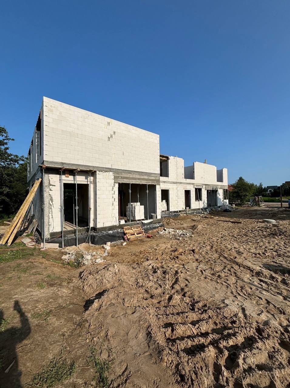 Wznoszenie ścian z białego bloczka na budowie domu jednorodzinnego, widoczne zbrojenia i brakujące nadproża okienne, teren budowy z piaskiem.