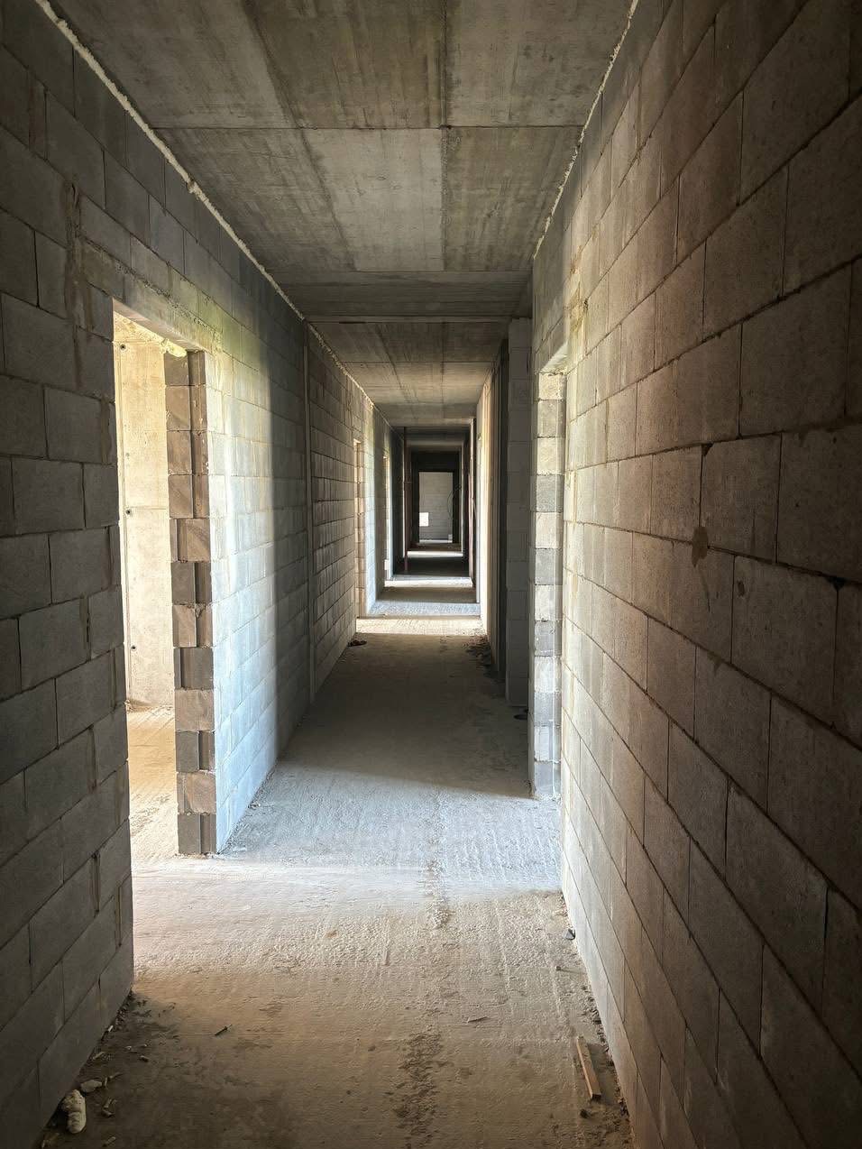 Wnętrze surowego budynku w trakcie budowy. Widok korytarza z szarymi ścianami z bloków i betonowym sufitem. Otwory drzwiowe wzdłuż ścian.