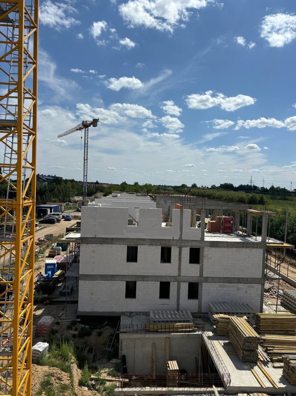 Wysoki kadr: budowa dwukondygnacyjnego budynku z białych bloczków, widoczny żuraw budowlany i częściowo żółty dźwig. Błękitne niebo z białymi obłokami w tle.