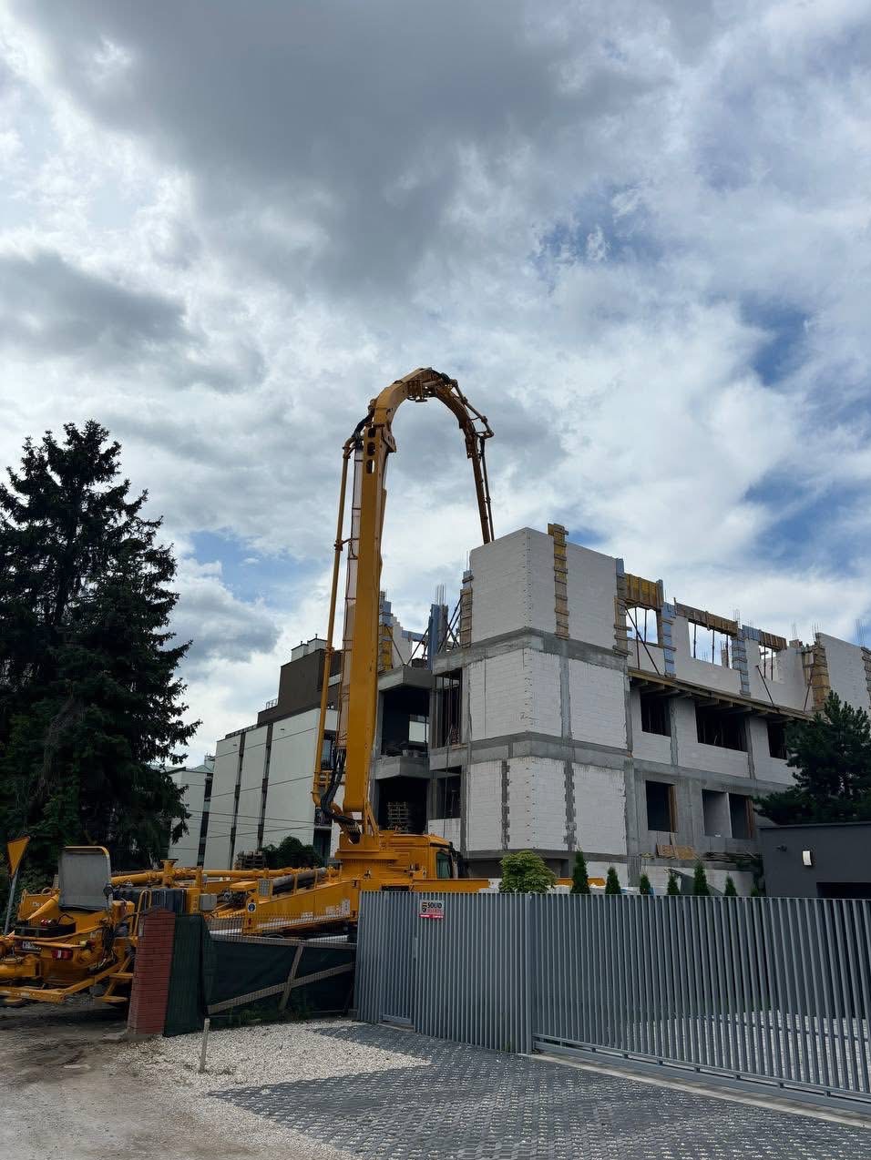 Budowa bloku z betonu komórkowego: żółta pompa do betonu podaje mieszankę na ściany, widoczne zbrojenia i szalunki okienne, pochmurne niebo w tle.