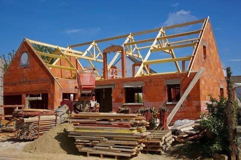 Surowy stan domu z czerwonej cegły z drewnianą konstrukcją dachu w trakcie budowy, z widocznym stosem desek i betoniarką na pierwszym planie.