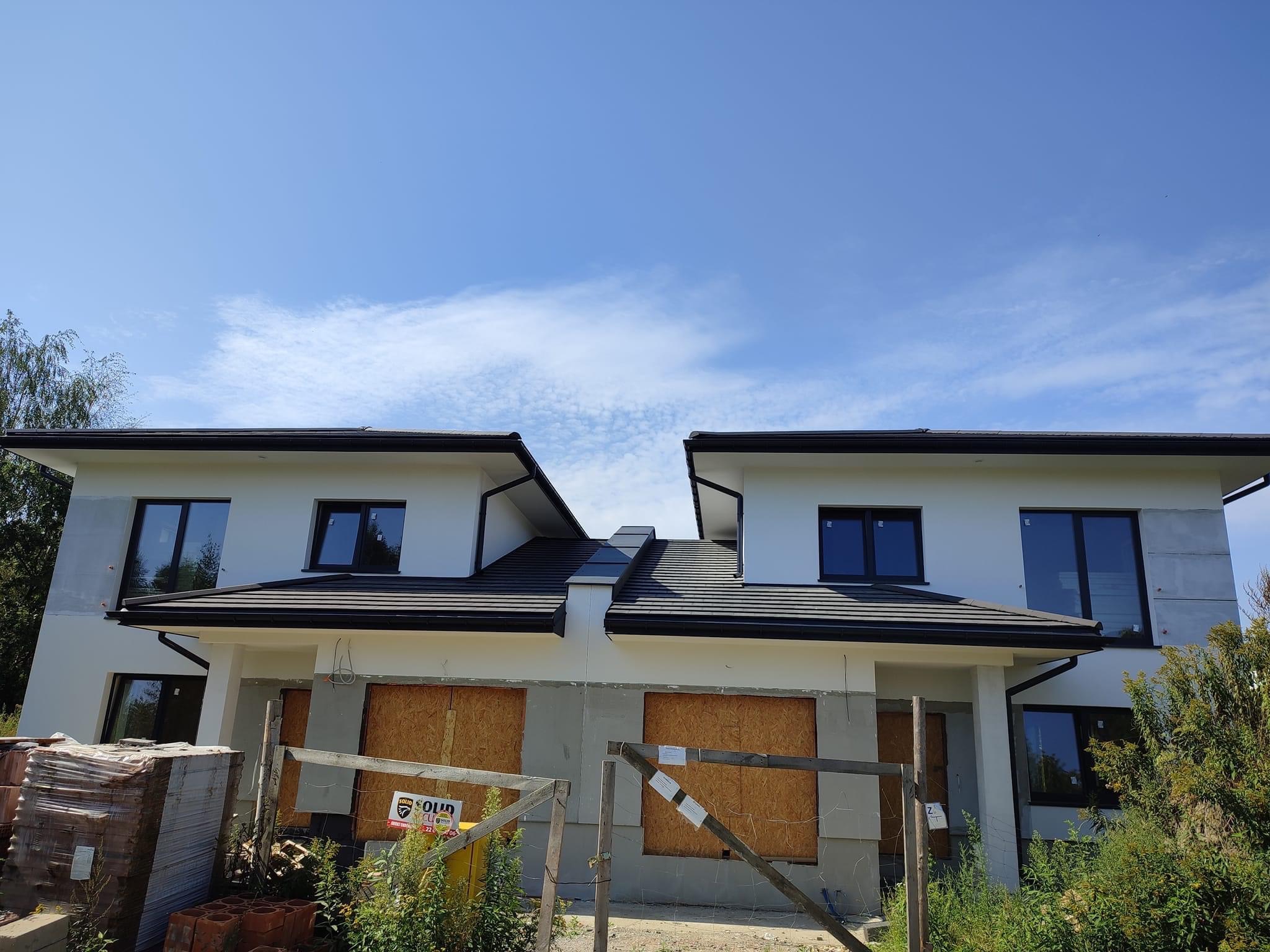 Bliźniak w stanie surowym zamkniętym z zabezpieczonymi oknami, widok z poziomu gruntu na tle błękitnego nieba.