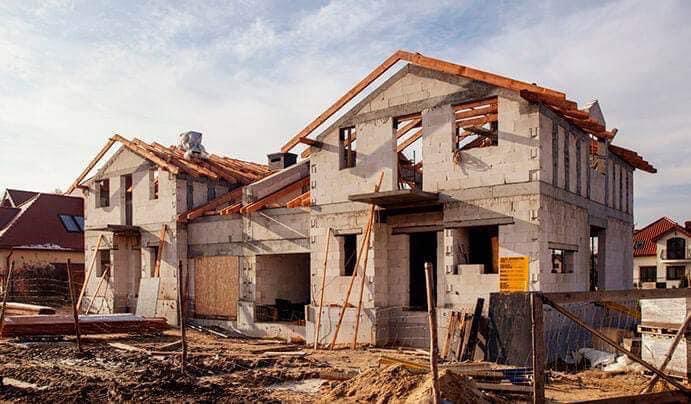 Dwa domy w trakcie budowy, ściany z betonu komórkowego, widoczne drewniane elementy konstrukcji dachu, teren budowy z ziemią i materiałami budowlanymi.