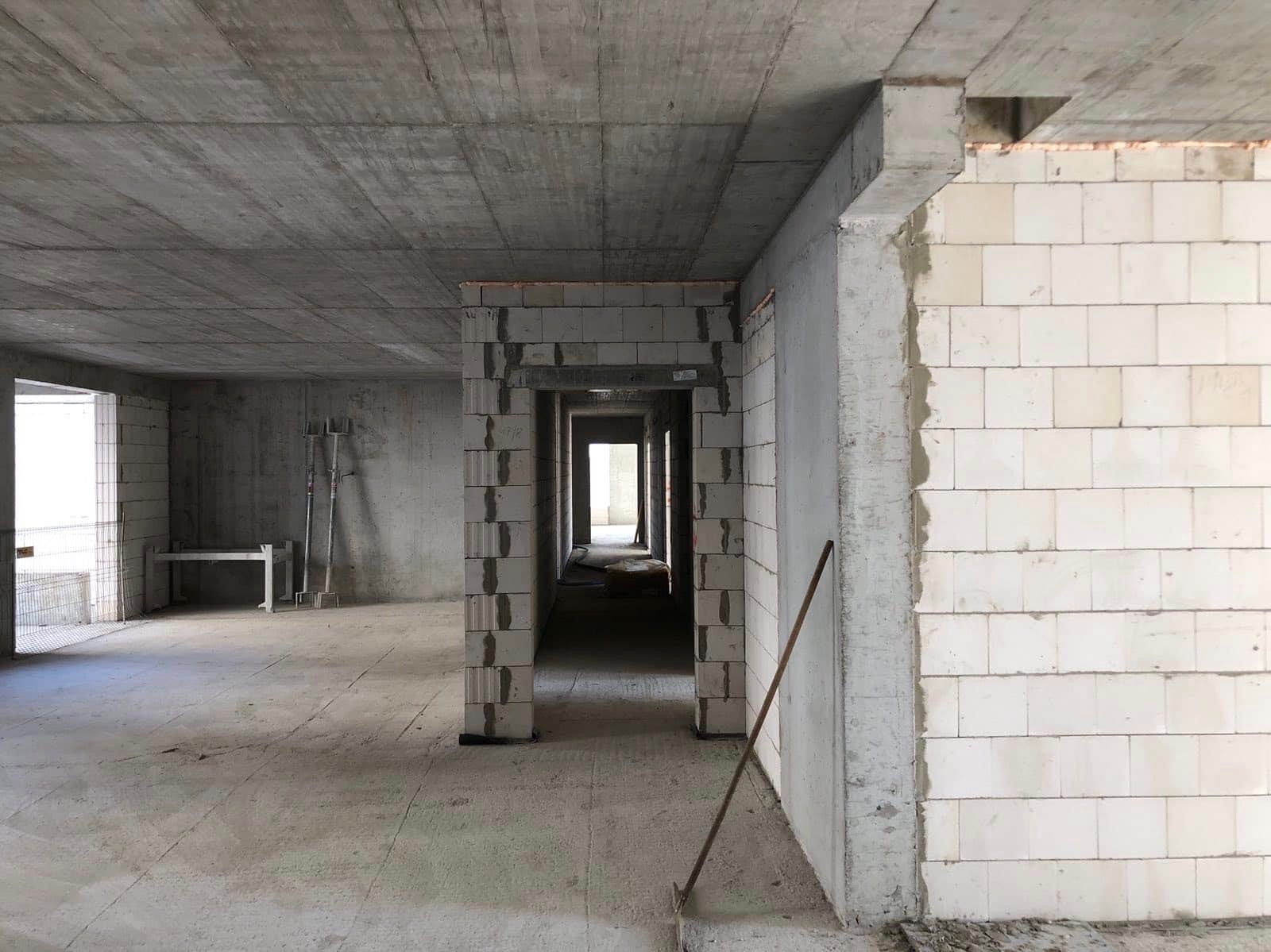 Surowy stan wnętrza domu w trakcie budowy: widoczne ściany z bloczków, betonowy strop, przygotowane otwory drzwiowe i prowizoryczne podpory.