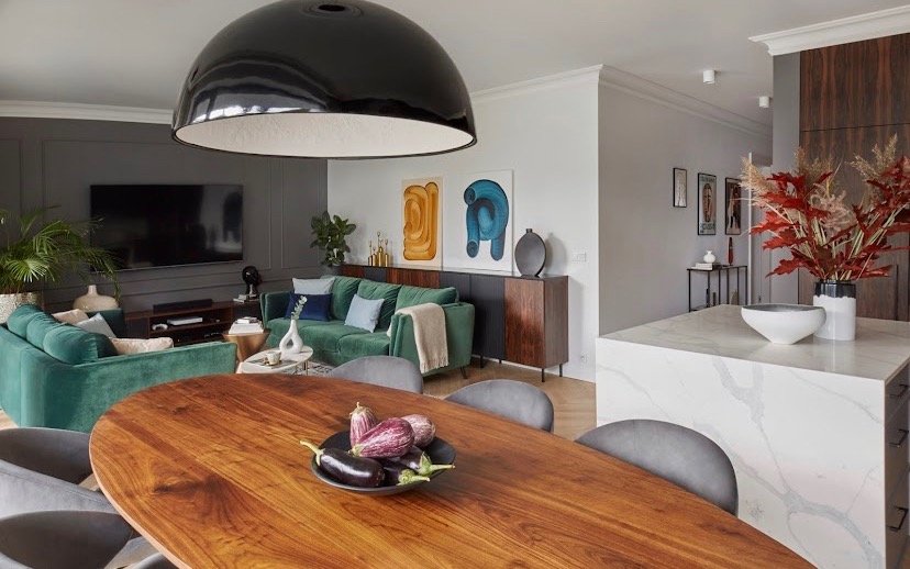 Nowoczesne wnętrze mieszkania z zieloną sofą, drewnianym stołem i dekoracjami, oświetlone dużą, czarną lampą wiszącą. Na stole leżą bakłażany w czarnej misce.