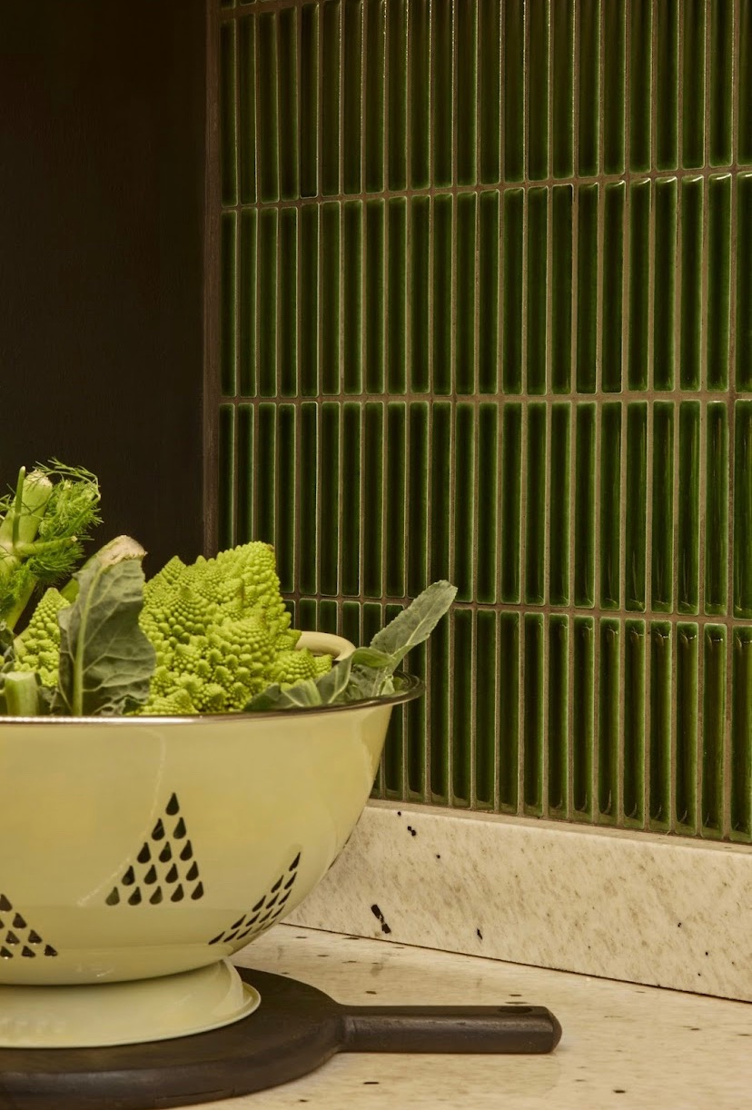 Zielona, wąska mozaika na ścianie w kuchni, obok durszlaka z romanesco i fenkułem na blacie.