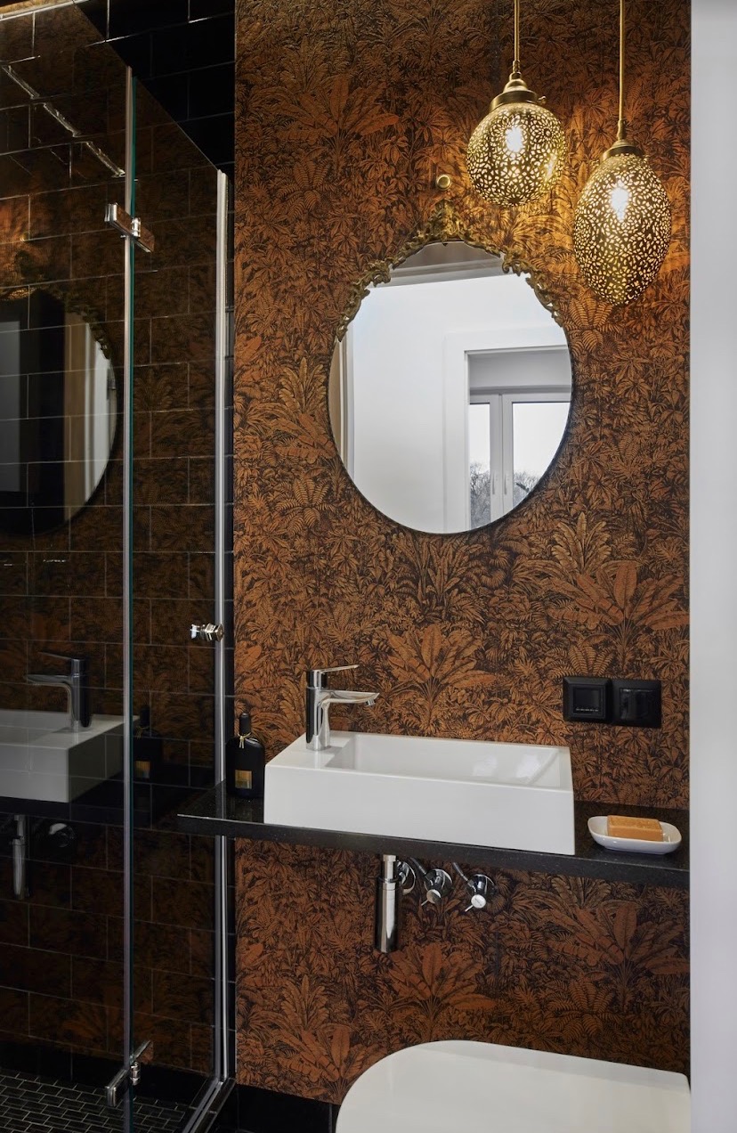 Eklektyczna łazienka z tapetą w motywy roślinne, okrągłym lustrem w złotej ramie, wiszącymi lampami ażurowymi i białą, prostokątną umywalką nablatową.