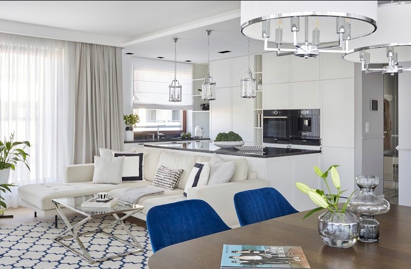 Jasne, przestronne wnętrze mieszkania z otwartą kuchnią, beżową sofą narożną, niebieskimi krzesłami i nowoczesnym oświetleniem wiszącym.