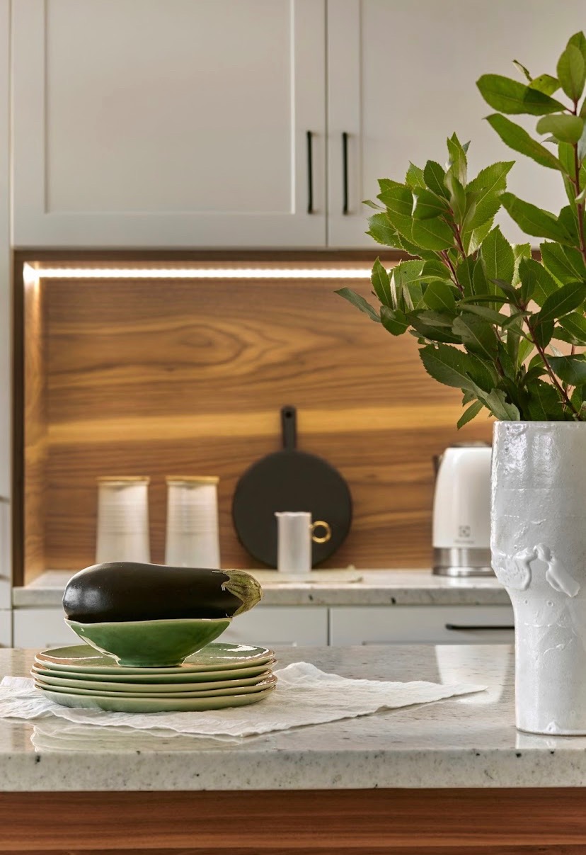 Nowoczesna kuchnia z blatem z jasnego kamienia, na którym leżą zielone talerze z bakłażanem, a w tle drewniana wnęka z oświetleniem LED i białymi szafkami.