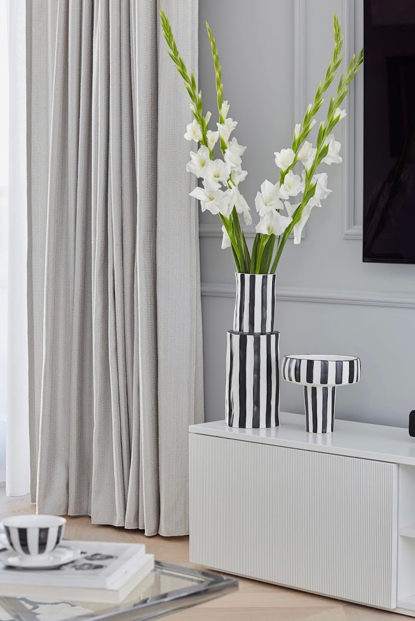 Minimalistyczny salon z szarymi zasłonami, białymi gladilolami w czarno-białych wazonach w paski na białej komodzie z ryflowanymi frontami.