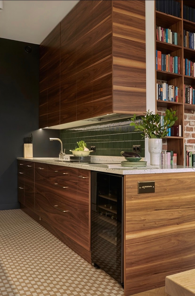 Nowoczesna kuchnia z drewnianymi szafkami o wyraźnym usłojeniu, zielonymi płytkami na ścianie, blatem z jasnego kamienia, wbudowaną chłodziarką do wina i regałem z książkami w tle.