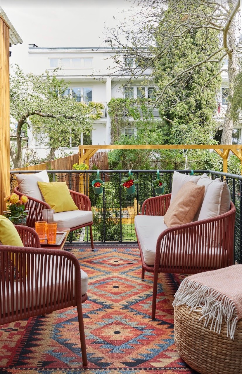 Przestronny balkon z kompletem wypoczynkowym z brązowej plecionki, wzorzystym dywanem w odcieniach czerwieni i niebieskiego, oraz widokiem na budynek mieszkalny i zieleń