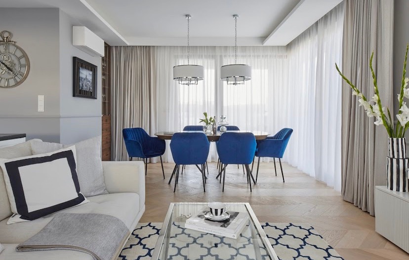 Salon z jadalnią: biała sofa z dekoracyjnymi poduszkami, szklany stolik kawowy z filiżanką, okrągły stół jadalny otoczony sześcioma granatowymi krzesłami, dwie lampy wiszące, jasne zasłony...
