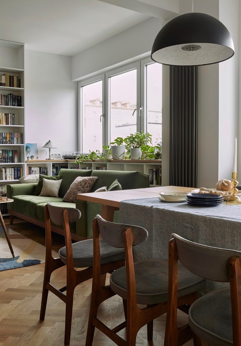 Jasne wnętrze pokoju dziennego z zieloną sofą, regałem z książkami, stołem z nakryciem i krzesłami w stylu retro, oświetlone wiszącą lampą z ażurowym wnętrzem
