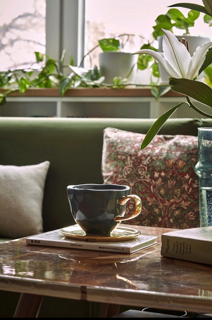 Szara, fakturowana filiżanka z złotym uchem na złotym spodku, ułożona na książce na błyszczącym, brązowym stoliku kawowym; w tle zielona kanapa, wzorzysta poduszka i okno z roślinami doniczkowymi.