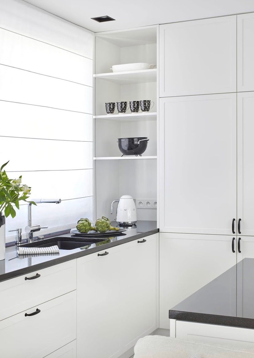 Jasna kuchnia w stylu skandynawskim: białe szafki z czarnymi uchwytami, czarny blat, zlewozmywak i armatura, na blacie dwa karczochy na czarnym talerzu, biały czajnik Smeg, na półkach czarne kubki...