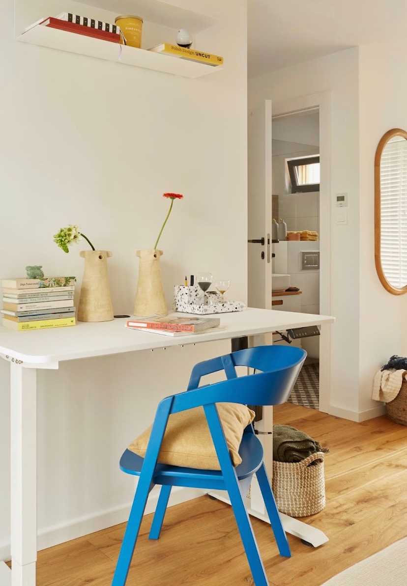 Jasne wnętrze z białym biurkiem, niebieskim krzesłem z poduszką, książkami, wazonami z kwiatami i widokiem na łazienkę w tle.