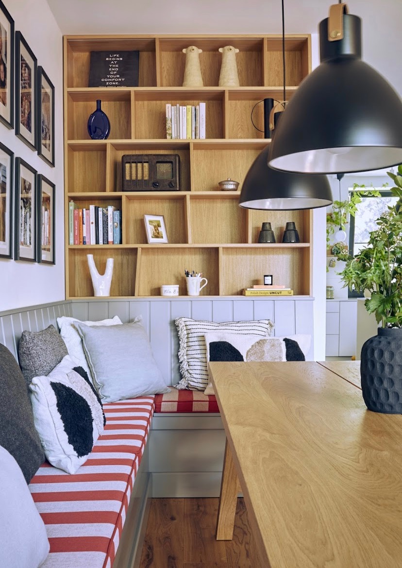 Drewniany regał z dekoracjami i książkami, narożna ławka z poduszkami w jadalni, oświetlona wiszącymi lampami.
