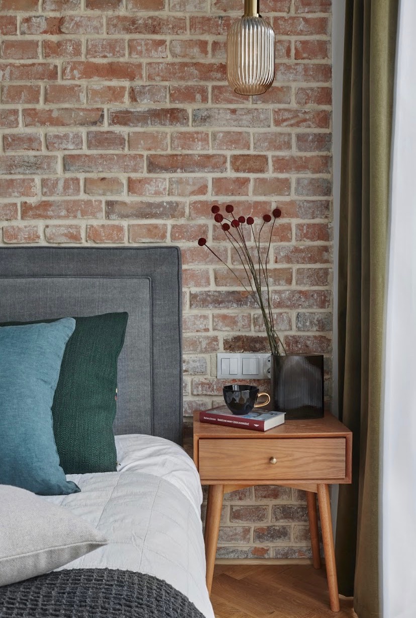 Sypialnia z ceglaną ścianą, drewnianą szafką nocną, szarą ramą łóżka i turkusowymi poduszkami, dekorowana wazonem z kulistymi kwiatami i czarnym kubkiem na książce.