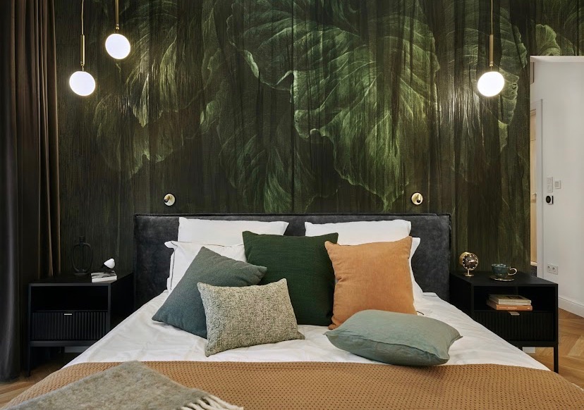 Sypialnia z tapetą w motywy tropikalnych liści, oświetlona wiszącymi lampami z kulistymi kloszami, z łóżkiem z szarym zagłówkiem i poduszkami w odcieniach zieleni i pomarańczu.
