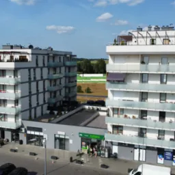 Nowoczesne apartamentowce z balkonami zabezpieczonymi szklanymi balustradami, widok z góry na osiedle z sklepami w parterze i wiatrakiem w tle.