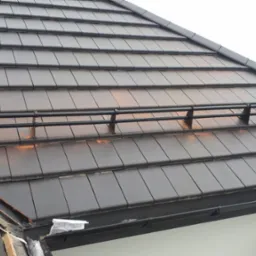 Ciemna dachówka ceramiczna z zamontowanym systemem zabezpieczającym przed osuwaniem się śniegu, widoczne elementy mocujące, fragment orynnowania i uszkodzone miejsce na styku połaci dachu.