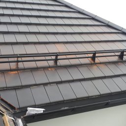 Pdn.drew - Ciemna dachówka ceramiczna z zamontowanymi czarnymi barierkami przeciwśniegowymi; w tle widoczne zielone drzewa.