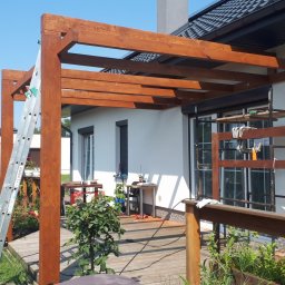 Pdn.drew - W trakcie budowy drewniana konstrukcja z belek na tarasie przylegająca do białego domu, widoczna drabina oparta o kolumnę i rusztowanie przy ścianie. Na pierwszym planie fragment drewnianej...