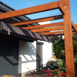 Pdn.drew - Drewniana konstrukcja z rozsuwanym, czarnym zadaszeniem, przylegająca do białego budynku z szarymi roletami, otoczona zielenią i czerwonymi kwiatami.