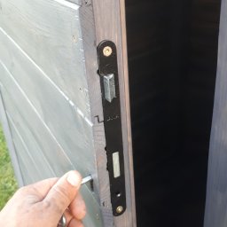 Pdn.drew - Otwieranie drewnianej altany pomalowanej na szaro z czarnym zamkiem, widoczny klucz w dłoni.