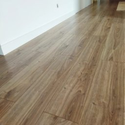 Pdn.drew - Ułożone panele podłogowe w odcieniach brązu z wyraźnym wzorem drewna, widoczne białe listwy przypodłogowe przy ścianie.