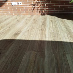 Pdn.drew - Jasne panele podłogowe ułożone przy ceglanej ścianie z widocznym gniazdkiem elektrycznym, oświetlone popołudniowym słońcem rzucającym cień liści.