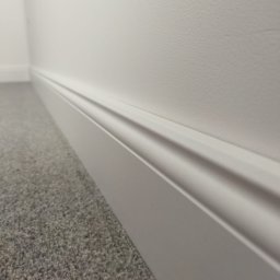 Pdn.drew - Szczegółowe ujęcie białej listwy przypodłogowej z delikatnym frezem, przylegającej do ściany i szarej wykładziny dywanowej, z widocznym narożnikiem ściany w tle.