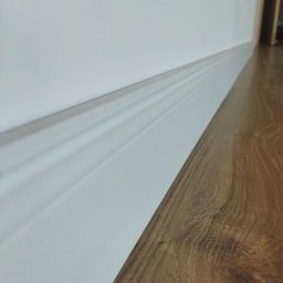 Pdn.drew - Detal drewnianej podłogi panelowej z białą, ozdobną listwą przypodłogową, widoczne łączenie paneli i subtelne wzory drewna.