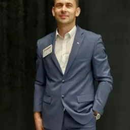 Mężczyzna w błękitnym garniturze, białej koszuli w drobne kropki i z identyfikatorem na piersi, pozujący na tle ciemnej zasłony. Widoczny zegarek na lewym nadgarstku.