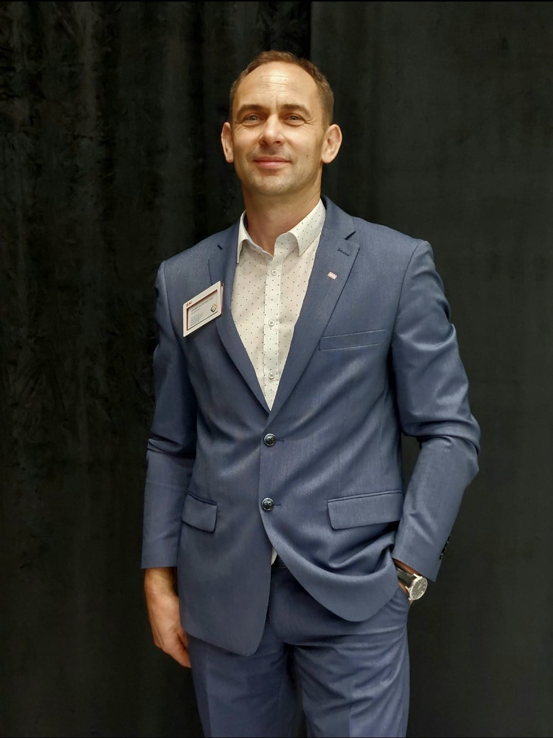 Mężczyzna w błękitnym garniturze, białej koszuli w drobne kropki i z identyfikatorem na piersi, pozujący na tle ciemnej zasłony. Widoczny zegarek na lewym nadgarstku.