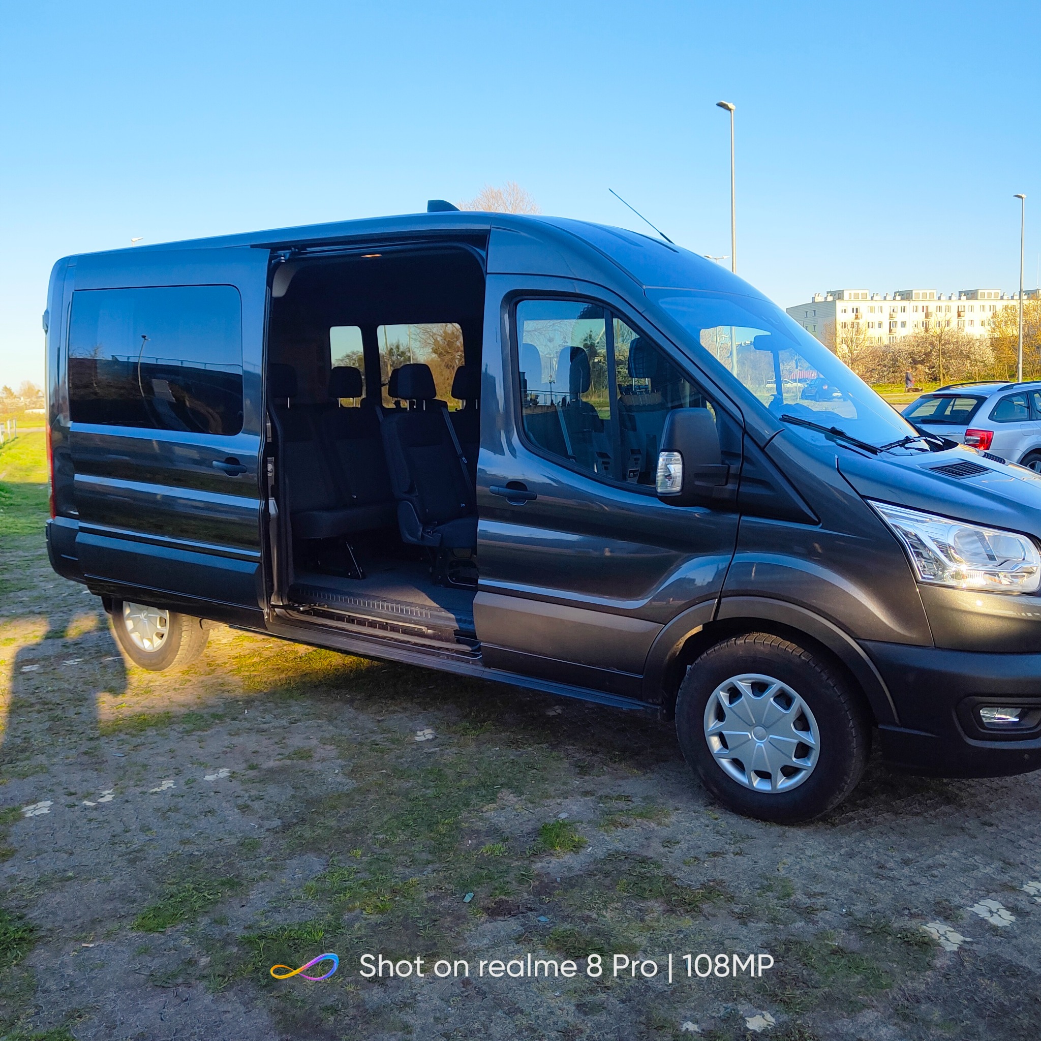 Szary bus z otwartymi drzwiami bocznymi, widoczne czarne fotele wewnątrz, na zewnątrz trawa i budynki w tle.