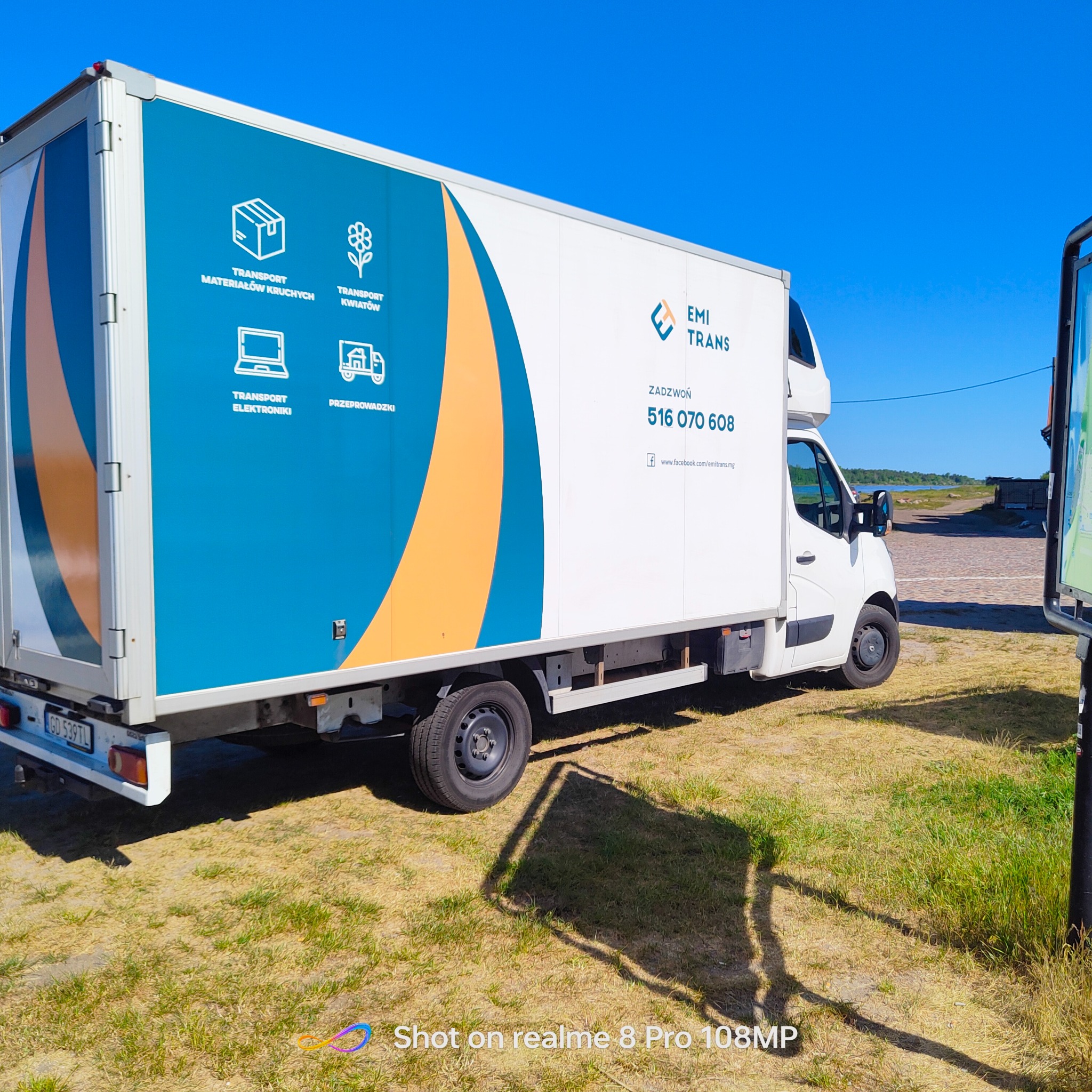 Biała furgonetka z niebiesko-pomarańczową grafiką informującą o transporcie materiałów kruchych, kwiatów, elektroniki i przeprowadzkach, zaparkowana na trawie w słoneczny dzień.