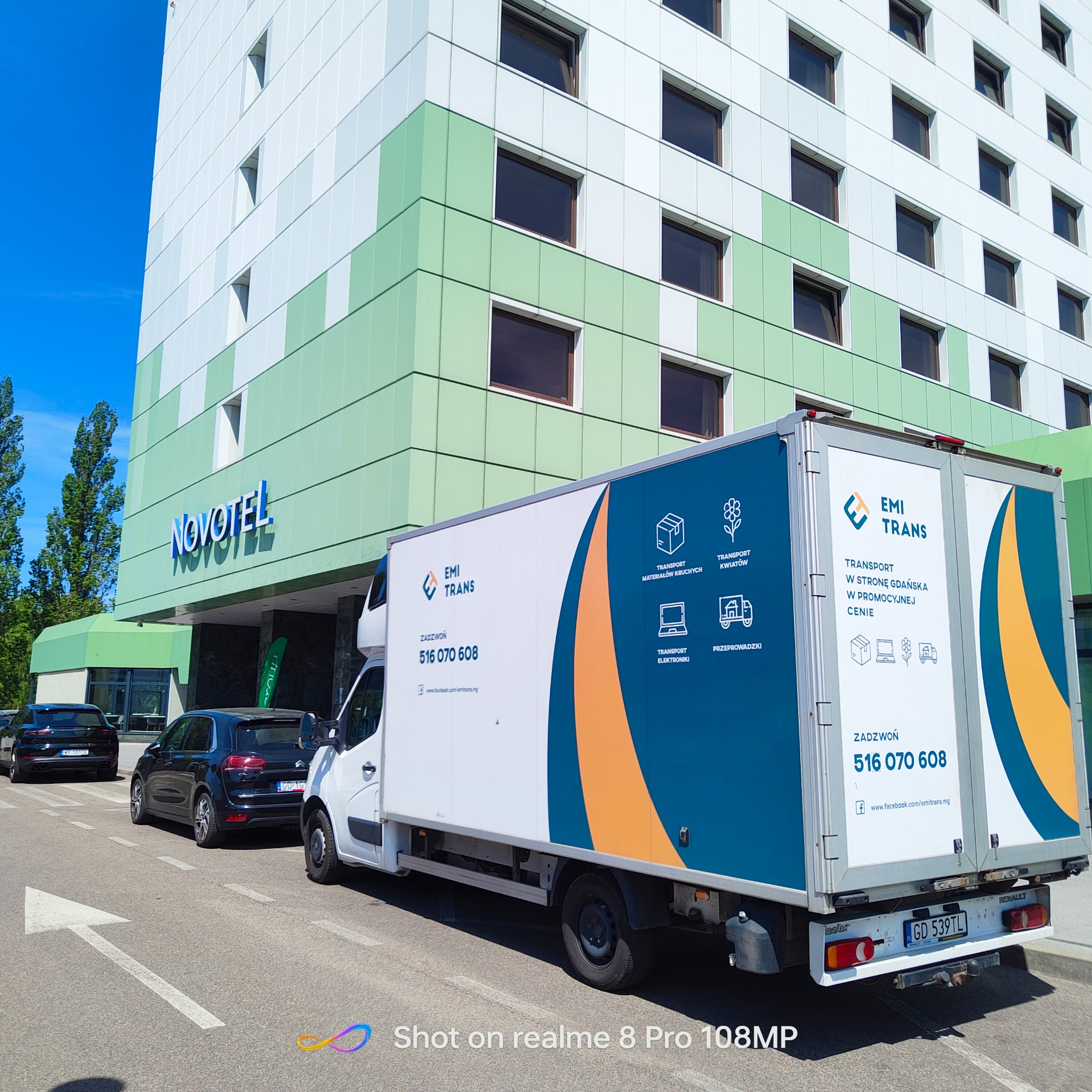 Biała furgonetka firmy EMI TRANS z niebiesko-pomarańczowym brandingiem zaparkowana przed hotelem Novotel, z widocznymi ikonami transportu materiałów kruchych, kwiatów, elektroniki i przeprowadzek.