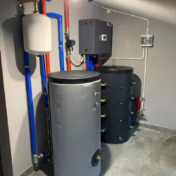 Dwa zbiorniki buforowe, szary i czarny, z podłączonymi rurami w kolorze czerwonym i niebieskim, zamontowane w rogu pomieszczenia z szarą podłogą i ścianami. Widoczny również biały zbiornik...