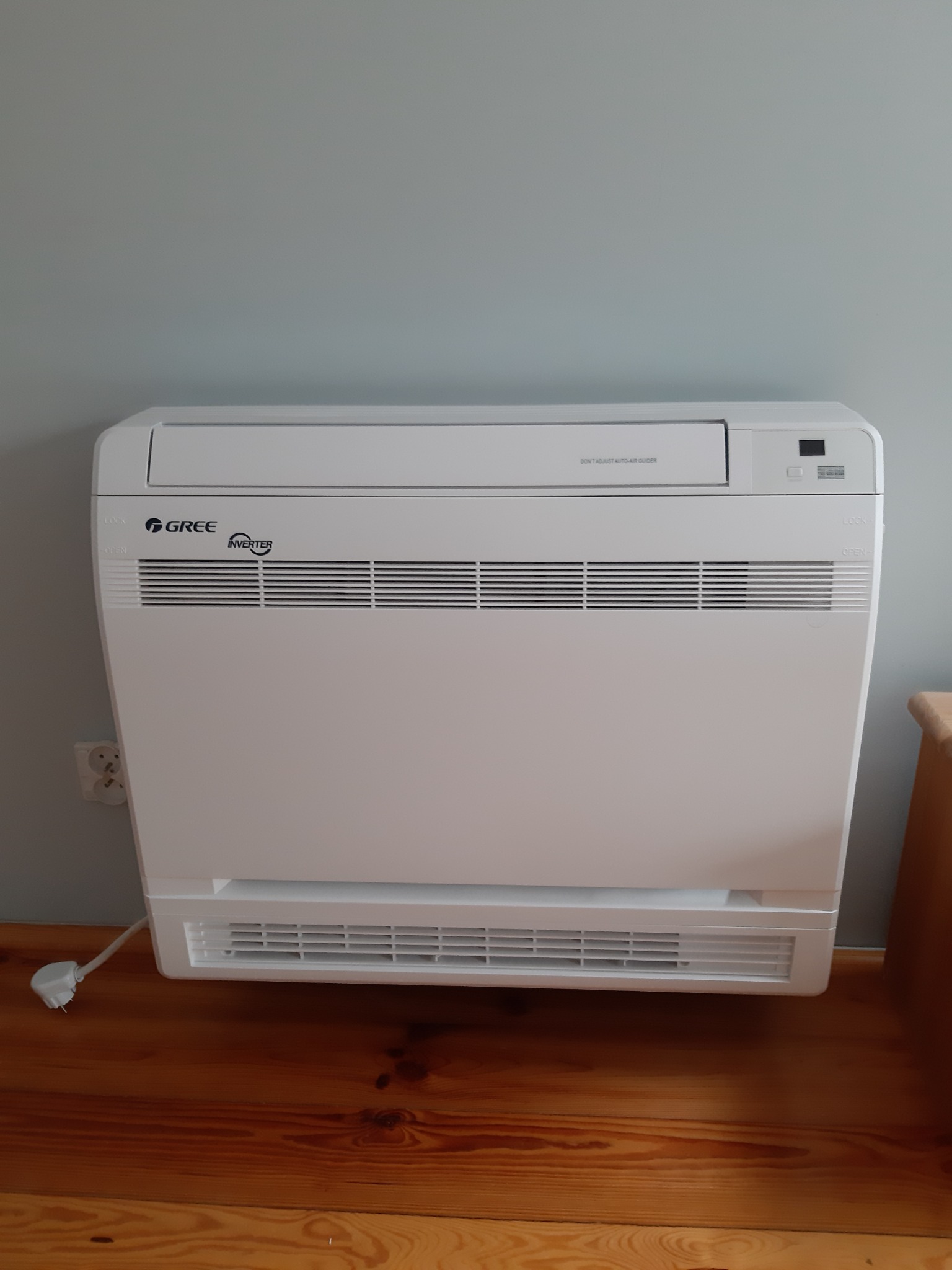 Biała, podłogowa jednostka klimatyzacji Gree Inverter, ustawiona na drewnianej podłodze, z widocznym kablem zasilającym i gniazdkiem elektrycznym.