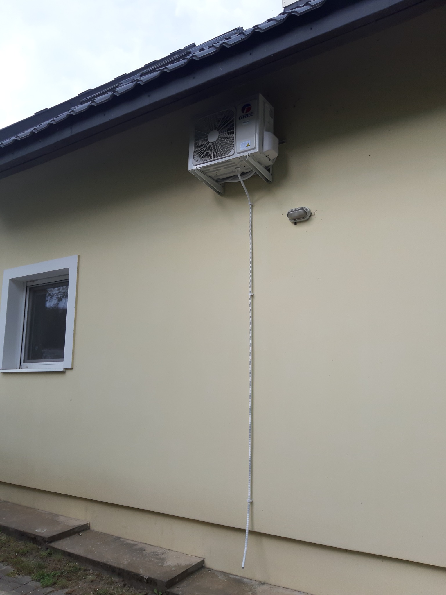 Zewnętrzna jednostka klimatyzacji marki Gree zamontowana na elewacji budynku z kremową ścianą i ciemnym dachem, widoczne białe rury prowadzące w dół ściany.