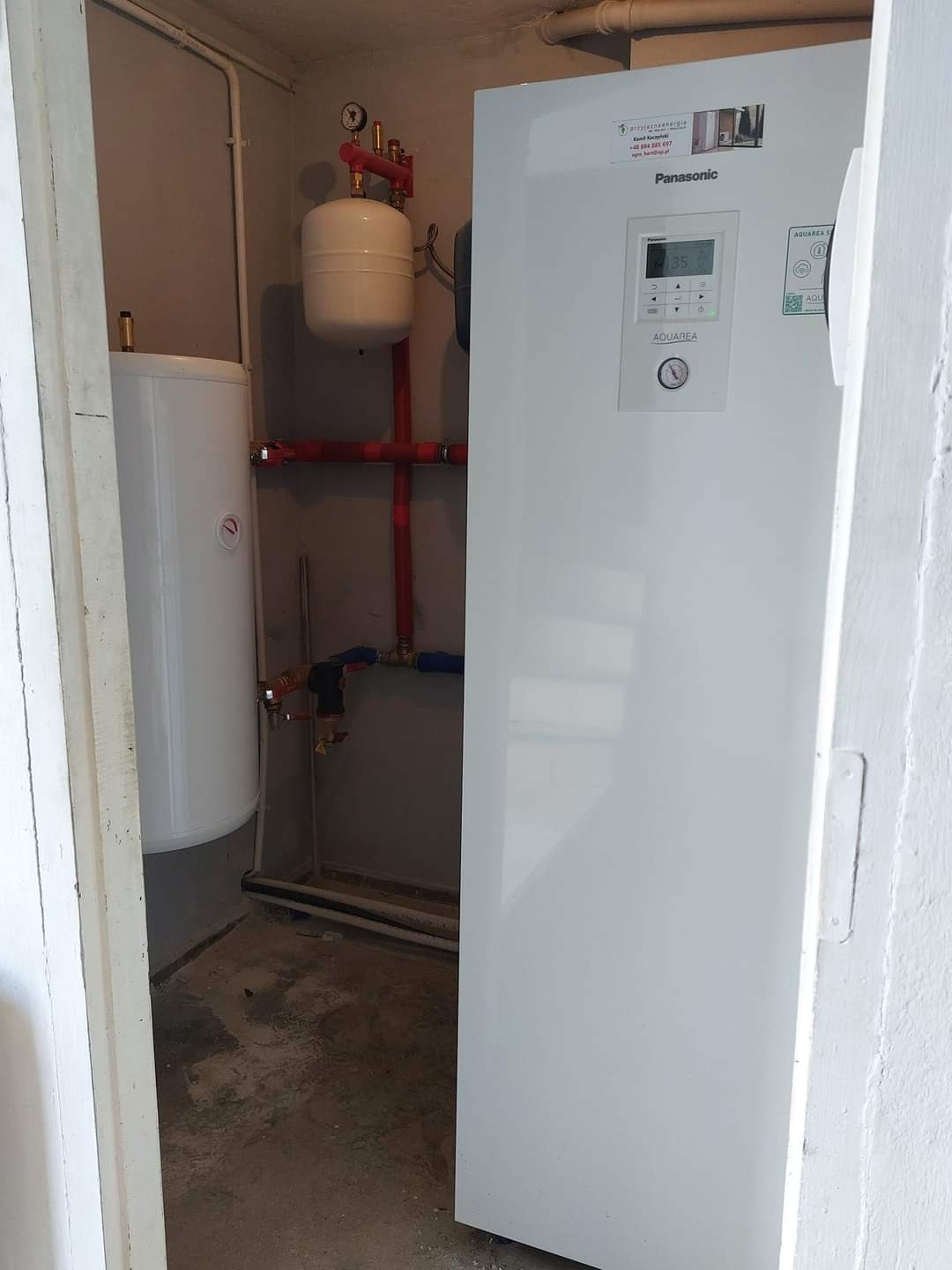 Zainstalowana pompa ciepła Panasonic Aquarea All In One o mocy 9 kW z wbudowanym zbiornikiem CWU 185 litrów i buforem 100 litrów, widoczna w kotłowni z szarymi ścianami i elementami instalacji...