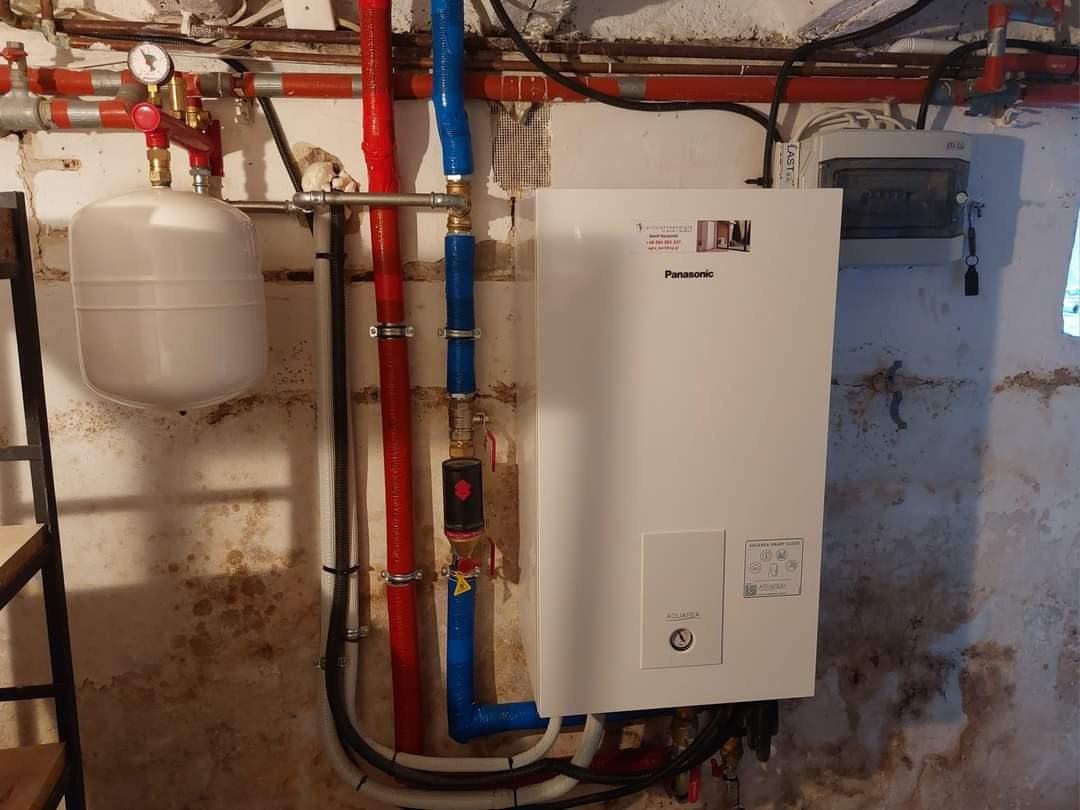 Biała jednostka wewnętrzna pompy ciepła Panasonic Aquarea typu split 9 kW T-CAP zamontowana na ścianie piwnicy z widocznymi rurami w kolorze czerwonym i niebieskim oraz zbiornikiem wyrównawczym.