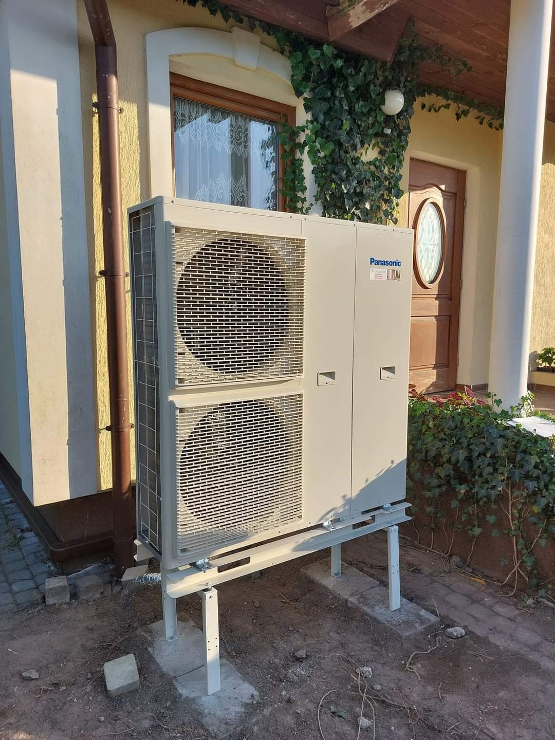 Zewnętrzna jednostka klimatyzacji typu split marki Panasonic zamontowana na metalowej konstrukcji przy ścianie budynku z brązowymi drzwiami i oknem porośniętym bluszczem.