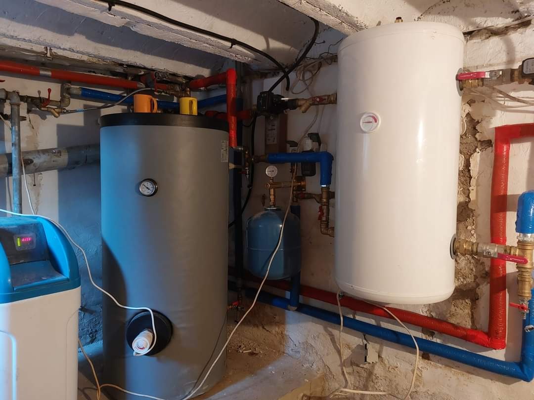 Dwa zbiorniki akumulacyjne ciepła, szary i biały, z widocznymi rurami instalacji grzewczej w pomieszczeniu technicznym z surowym wykończeniem ścian.